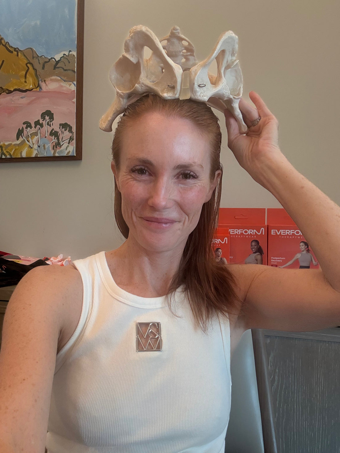 Rosie Dumbrell sitting at her desk smiling with an anatomical model of a pelvis on her head. 
